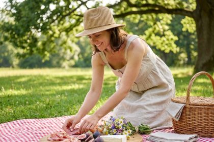 Femme élégante en robe de lin préparant un plateau de charcuterie et fromages sur une couverture à carreaux lors d'un pique-nique chic en plein air